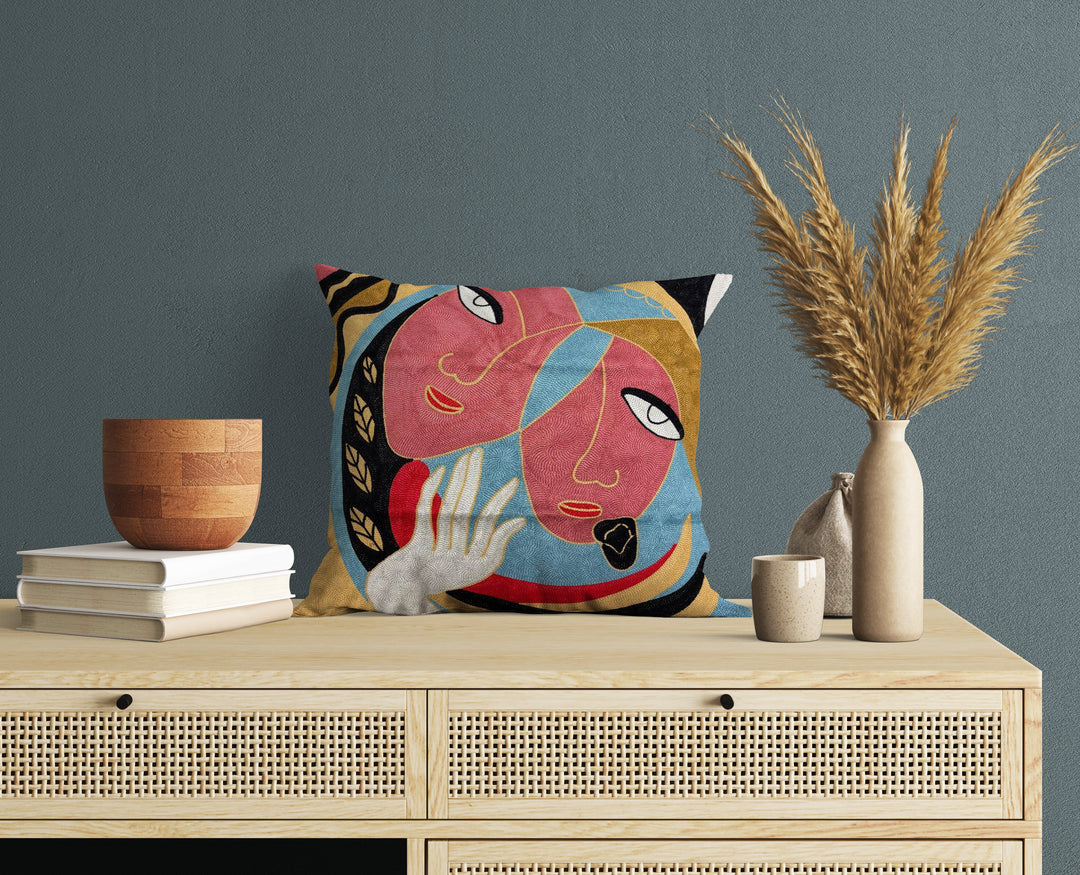 A cushion cover featuring an abstract design with two faces, displayed on a wooden table with decorative items around it.
