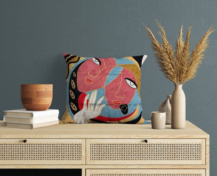 A cushion cover featuring an abstract design with two faces, displayed on a wooden table with decorative items around it.
