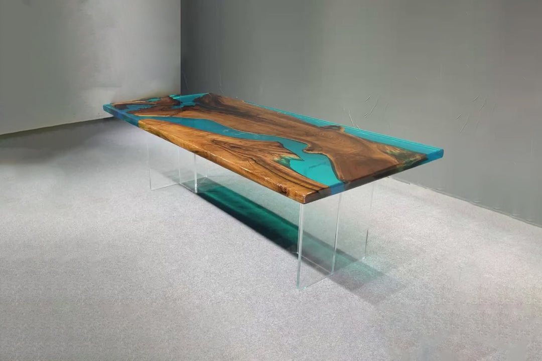 A rectangular dining table with a walnut wood tabletop and a blue epoxy resin river design, supported by a clear acrylic base.