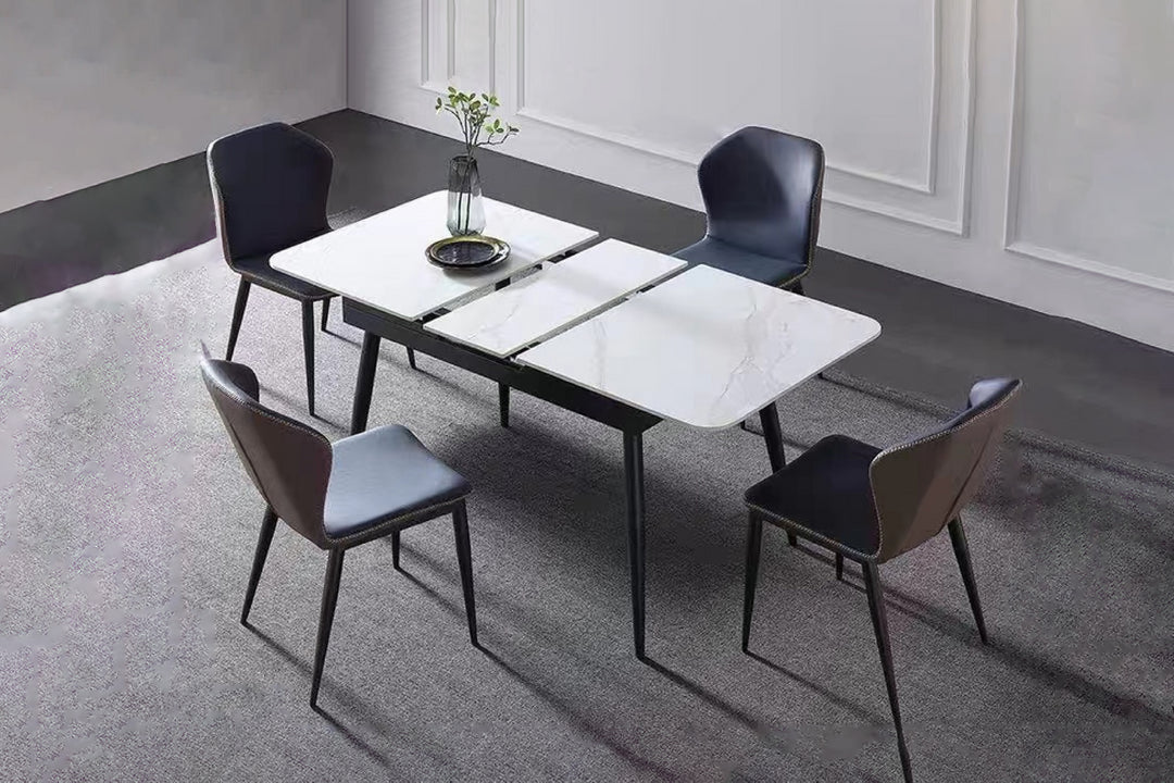 An extendable dining table with a white sintered stone top and black metal legs, surrounded by four modern chairs with gray and black upholstery. The table is set in a contemporary room with a minimalist decor.