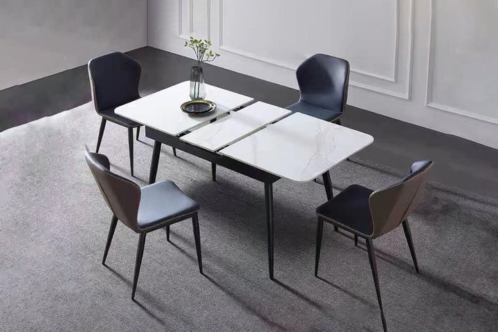 An extendable dining table with a white sintered stone top and black metal legs, surrounded by four modern chairs with gray and black upholstery. The table is set in a contemporary room with a minimalist decor.