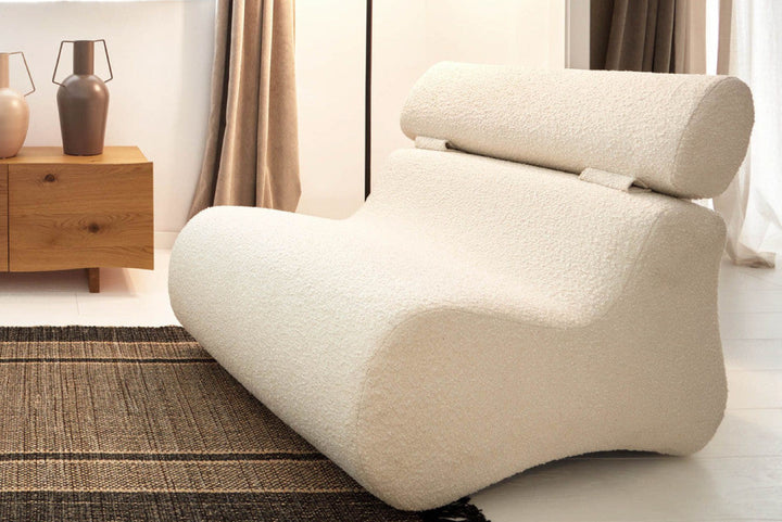 Modern beige armchair in a living room setting with wooden side table and curtains.