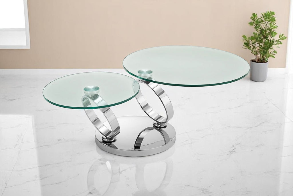 A swivel glass coffee table with a tempered glass top and a stainless steel base in a chrome finish, displayed on a white marble floor with a potted plant in the background.