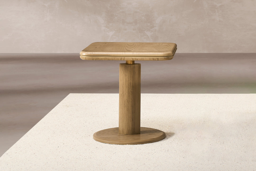 A square-shaped Bella Tavola side table with a light brown ash veneer finish, cylindrical leg, and round corners, placed on a white surface.