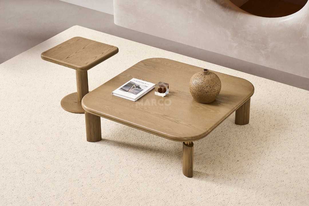 A low square coffee table made of ash veneer with round corners and stainless steel joints, accompanied by a round side table. The coffee table is set on a carpeted floor and has decorative items on it.