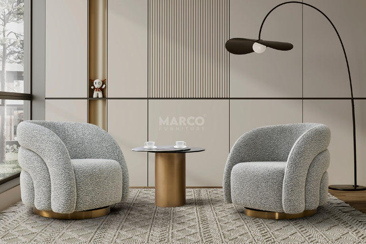The image features a modern interior setting with the Capri End Table at its center. The table has a round, cylindrical metal base with a gold finish, supporting a grey glass extension topped with a Chanel sintered stone surface.