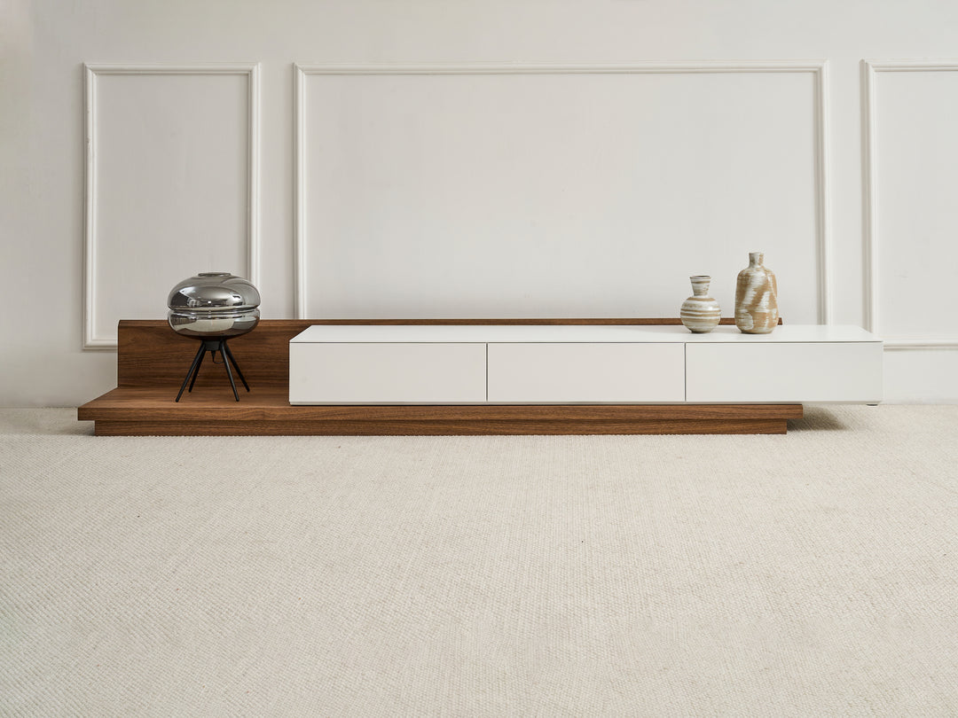 A contemporary extendable TV unit with a white top and natural walnut veneer base, featuring three drawers and a hole for cable management. It is displayed in a retracted state with decorative items on top.