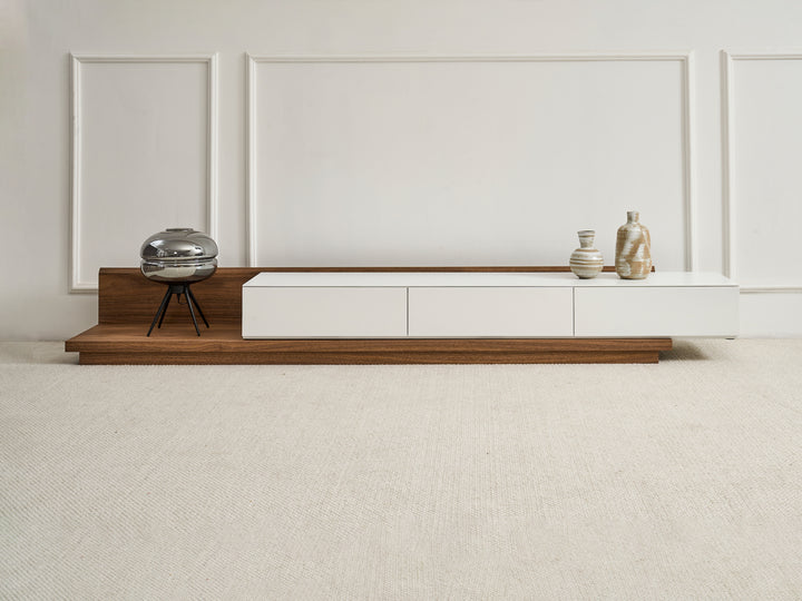 A contemporary extendable TV unit with a white top and natural walnut veneer base, featuring three drawers and a hole for cable management. It is displayed in a retracted state with decorative items on top.
