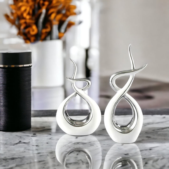 Two ceramic flame-shaped sculptures on a reflective surface, primarily white with silver accents, with a blurred background featuring a vase with flowers.