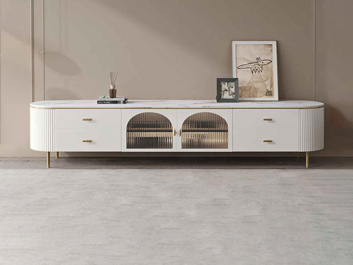 A modern white ceramic top TV unit with gold stainless steel legs and drawers, featuring fluted detailing and glass doors.