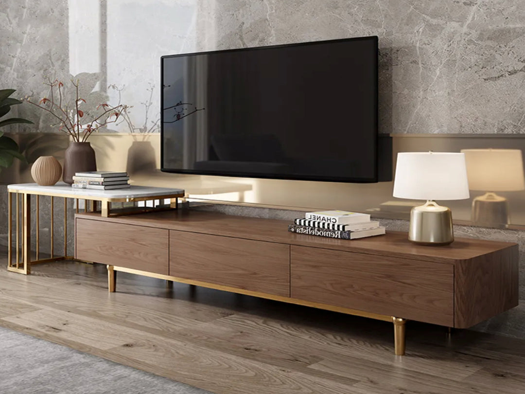 A modern walnut veneer TV unit with gold stainless steel legs, featuring three soft-close drawers and an extendable section with a sintered stone top. The TV unit is placed in a room with a flat-screen TV on top, flanked by two lamps on either side.