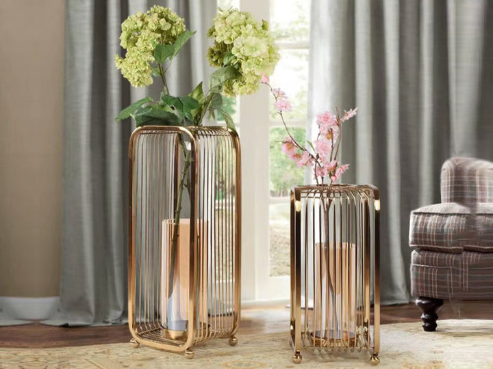 A set of two gold cage candle holders with clear glass inserts, one holding a bouquet of flowers, placed on a rug in a cozy room setting.