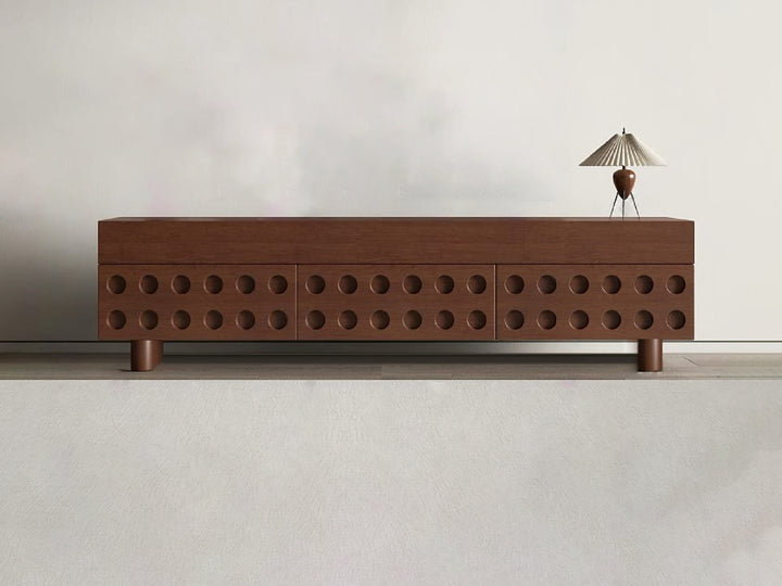 A modern TV cabinet with storage, featuring a unique circular cut-out design on the drawers, with a walnut finish and timber wood legs. A lamp is placed on top of the cabinet.