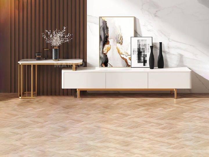 A modern white low-line TV unit with gold stainless steel legs and a marble extension, set against a wood panel wall with decorative items on top.