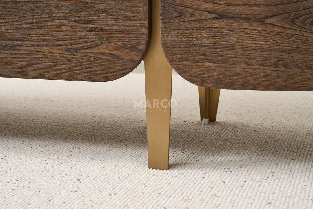 Close-up of a wooden chair leg with gold finish on a beige carpet, featuring the brand 'Marco'.