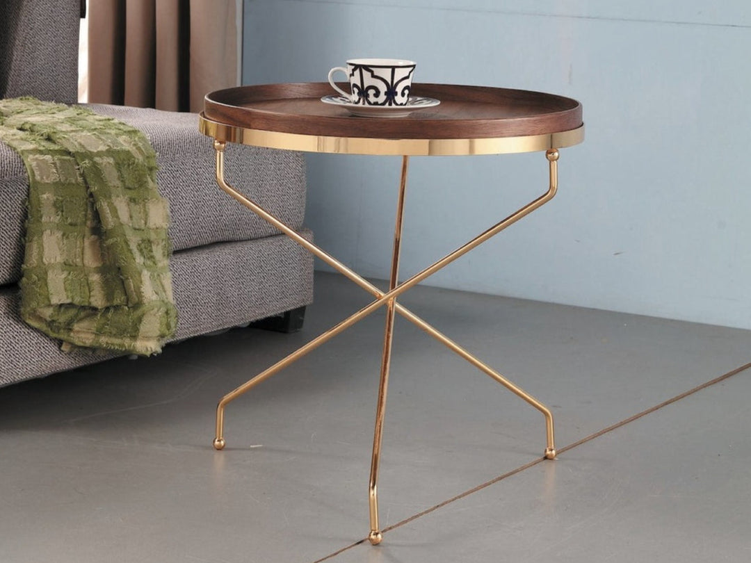 A modern round side table with a walnut veneer top and golden metal legs featuring an overlapping cross design, placed in a room with a sofa and a green checkered cloth.
