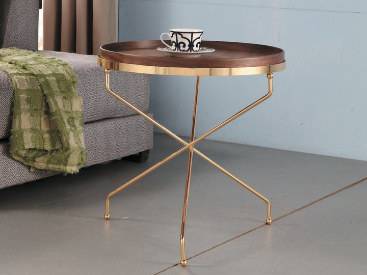 A modern round side table with a walnut veneer top and golden metal legs featuring an overlapping cross design, placed in a room with a sofa and a green checkered cloth.