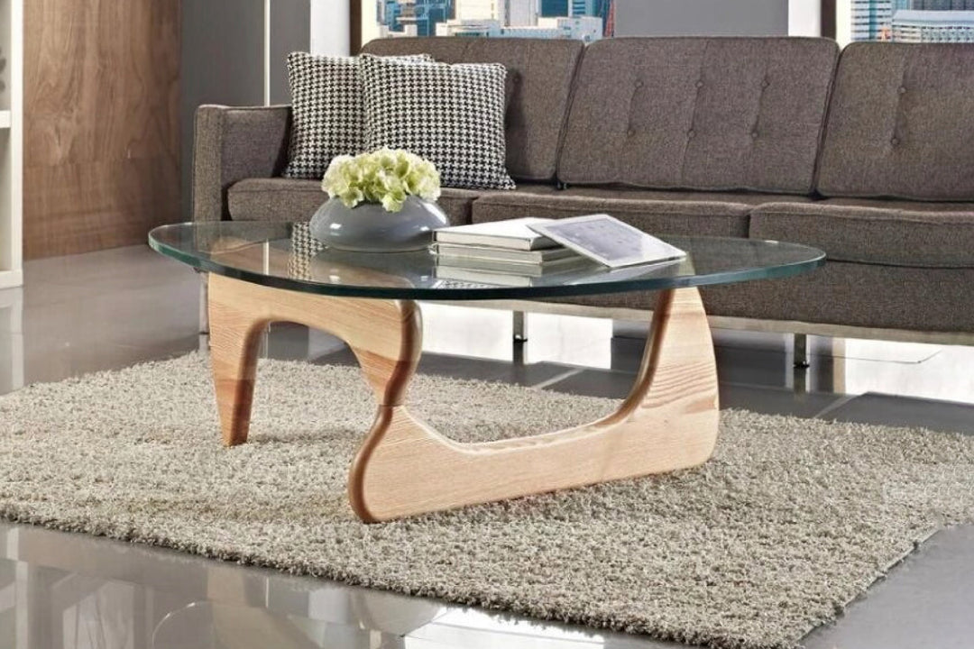 A Noguchi glass coffee table with ash wood legs on a carpet, with a glass top displaying a vase with flowers and a remote control.