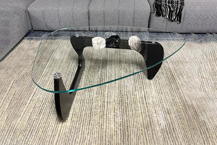 A Noguchi coffee table with a clear glass top and black wooden legs in an interlocking design, placed on a beige carpet.