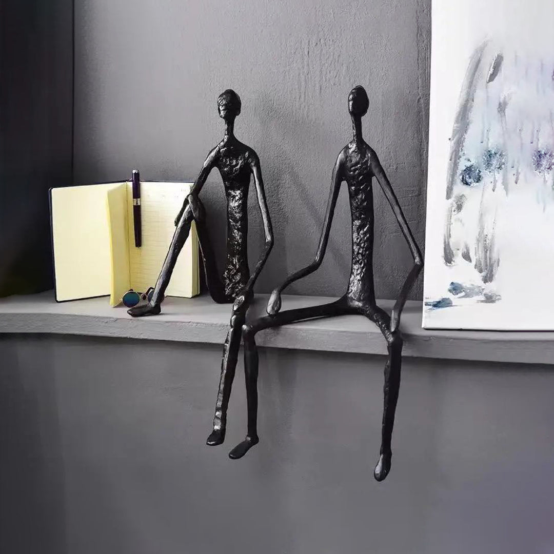 Two metallic sculptures of people on a shelf against a gray wall with a piece of abstract art.