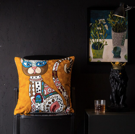 A cushion cover featuring a Picasso-inspired abstract design, predominantly orange with multiple geometric shapes and a stylized face, displayed in a dimly lit room with a framed picture and a lamp in the background.