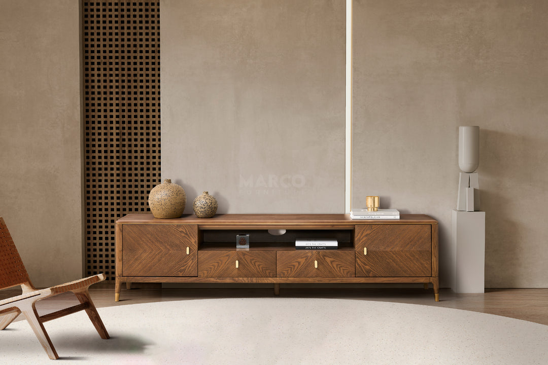 A modern walnut brown TV unit with storage drawers and cabinets, featuring brushed gold accents on short legs, placed against a concrete wall with decorative items on top.