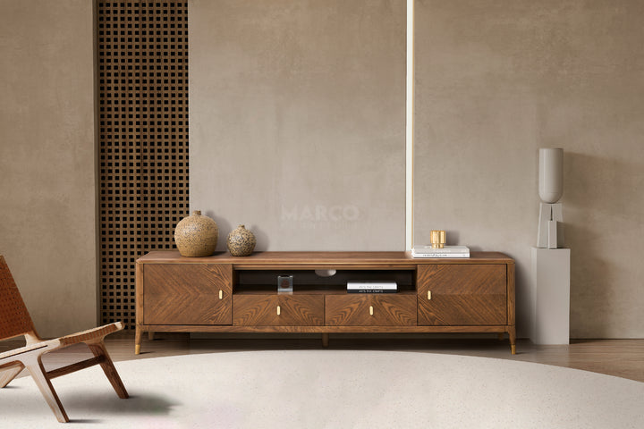 A modern walnut brown TV unit with storage drawers and cabinets, featuring brushed gold accents on short legs, placed against a concrete wall with decorative items on top.