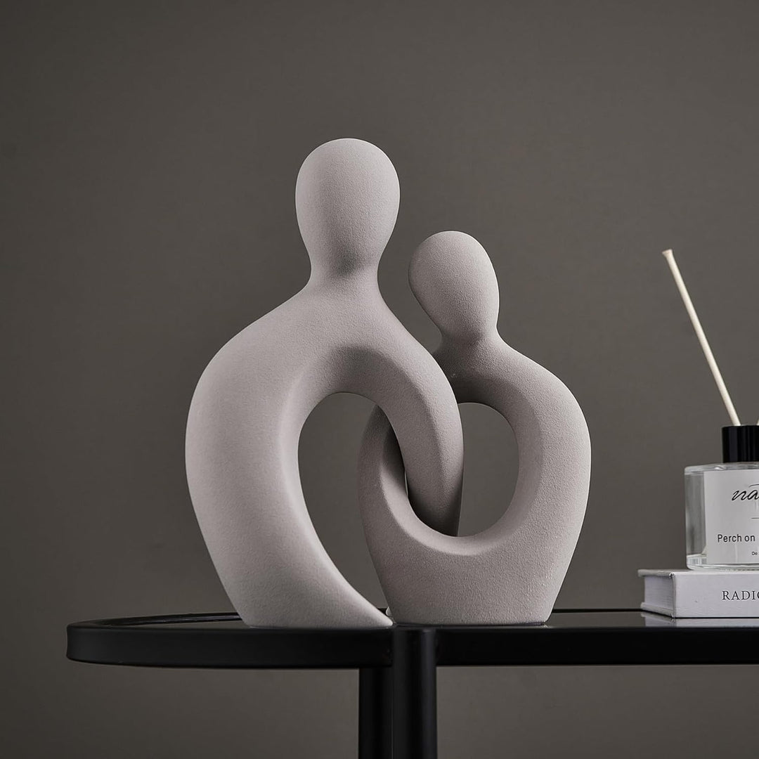 Two grey ceramic figurines of a romantic couple, intertwined in a heart-like shape, placed on a round table with a perfume bottle and a book in the background.