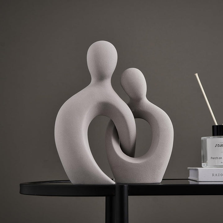 Two grey ceramic figurines of a romantic couple, intertwined in a heart-like shape, placed on a round table with a perfume bottle and a book in the background.