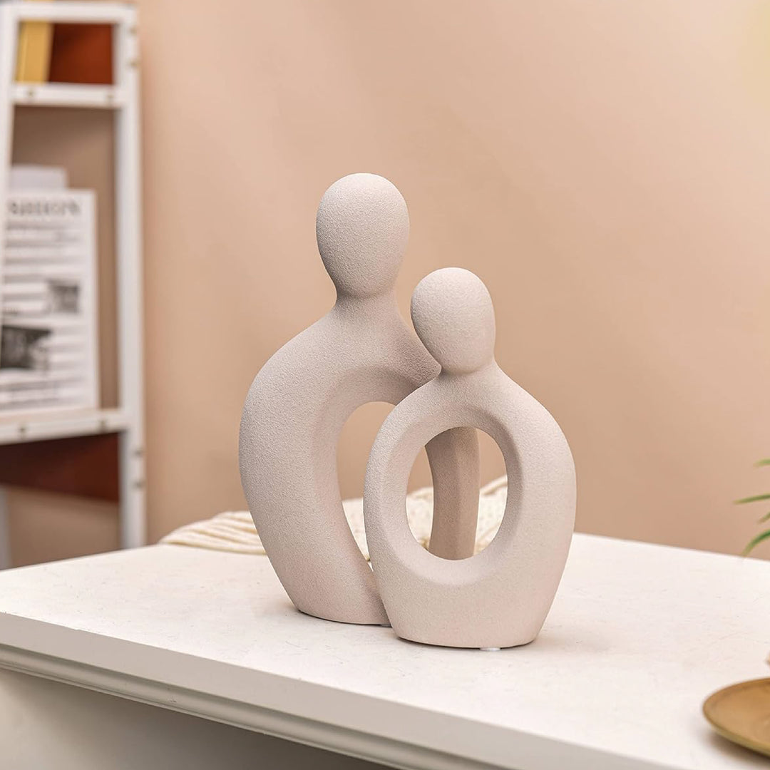 Ceramic sculpture of two abstract figures on a shelf against a beige wall.