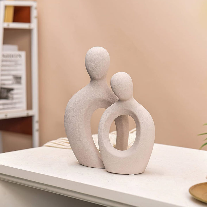 Ceramic sculpture of two abstract figures on a shelf against a beige wall.