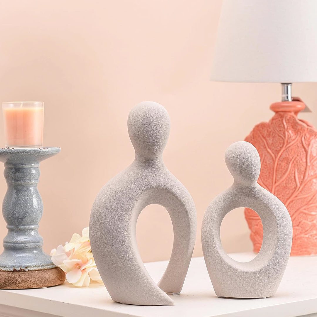 Two white abstract ceramic sculptures on a surface with a candle and decorative vase in the background.