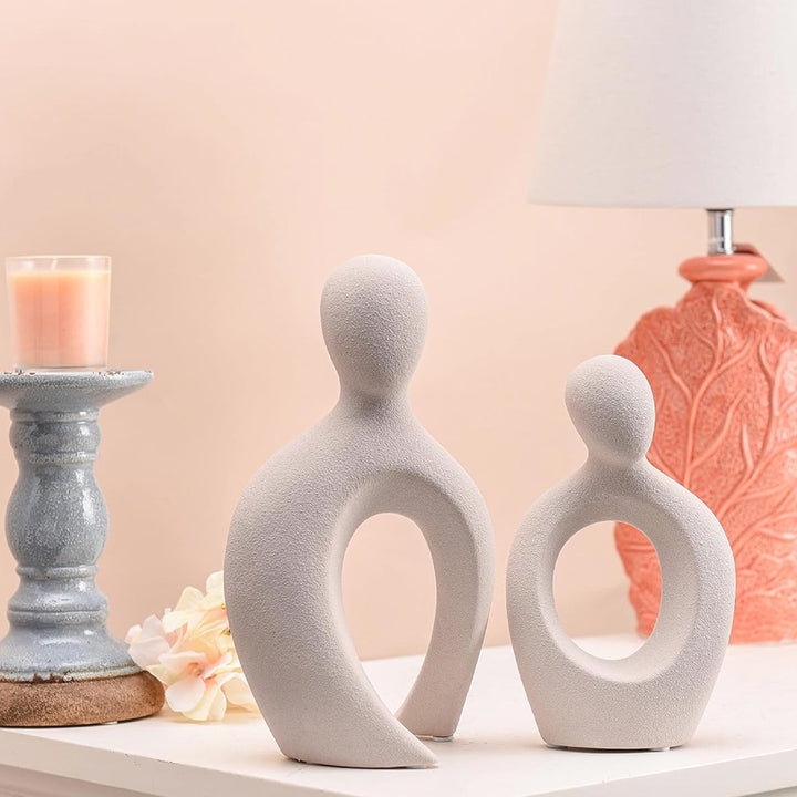 Two white abstract ceramic sculptures on a surface with a candle and decorative vase in the background.