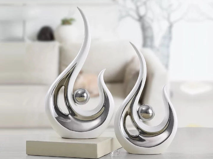 Two modern abstract ceramic phoenix figurines in white and silver colors, displayed on a shelf in a living room setting.