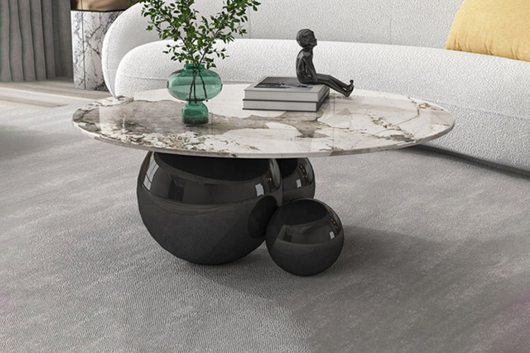 A round coffee table with a black metal base and a sintered stone top, placed on a grey floor with a white and beige pattern. The table has decorative items on it, including a plant pot and a sculpture.