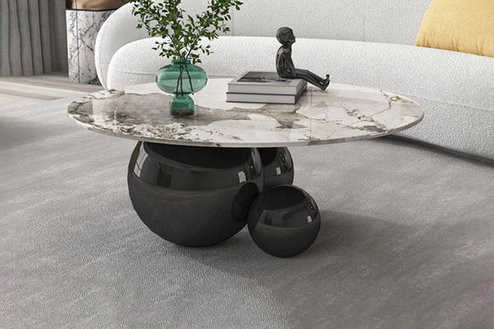 A round coffee table with a black metal base and a sintered stone top, placed on a grey floor with a white and beige pattern. The table has decorative items on it, including a plant pot and a sculpture.