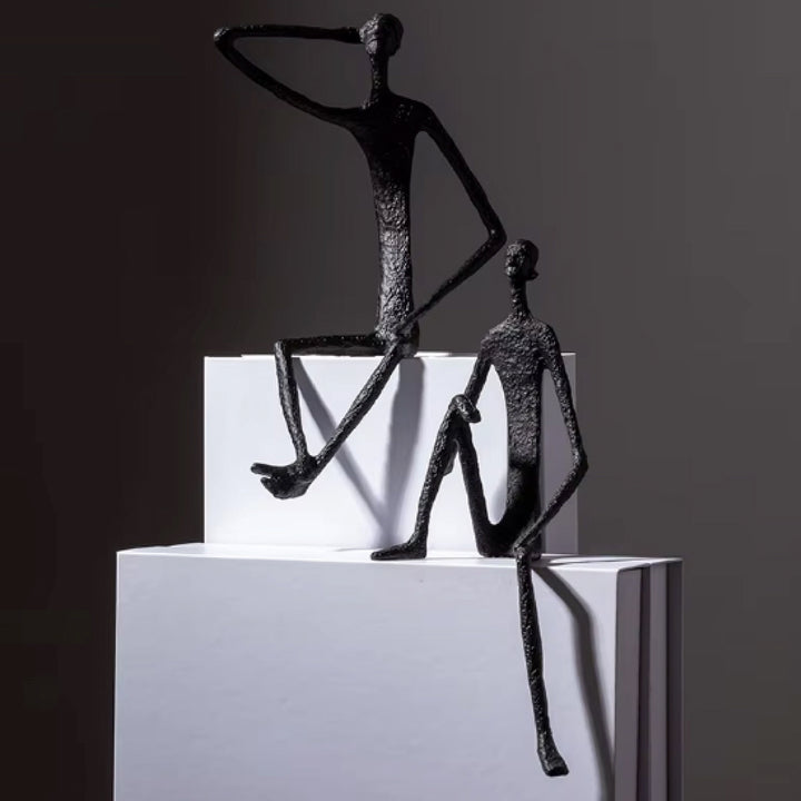 Abstract black sculpture of two figures on a white platform against a dark background