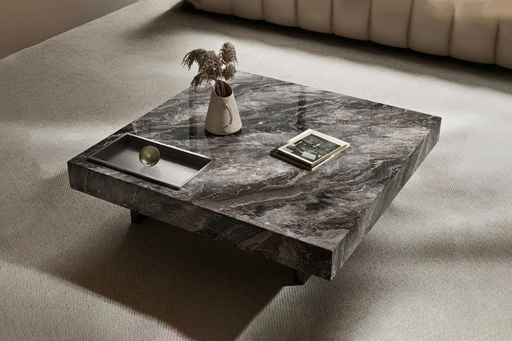 A natural marble coffee table with gray veining and a stainless steel base, accompanied by a metal tray with items on top, placed on a light-colored carpet.