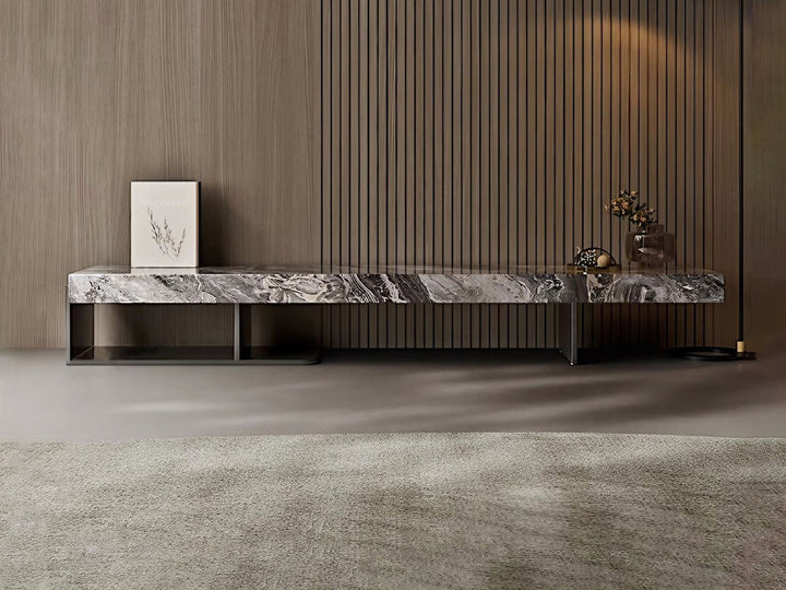 A modern marble top TV unit with a stainless steel base, placed against a dark paneled wall on a grey floor. The unit features a artwork frame and a decorative object on top.