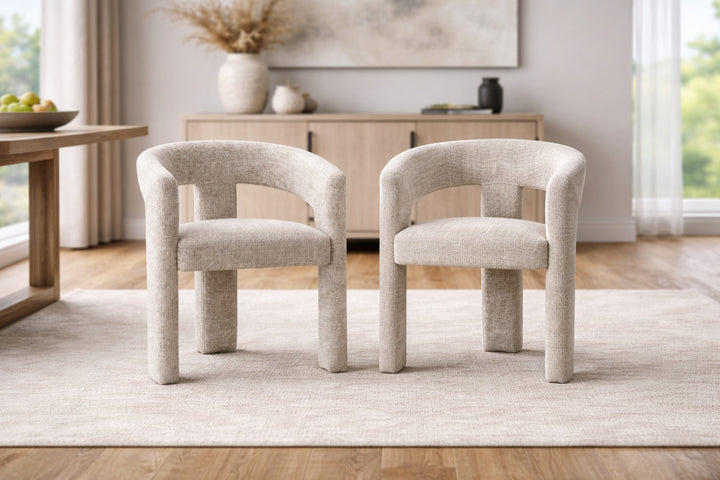 Two beige chairs on a light rug in a room with a wooden sideboard and window.