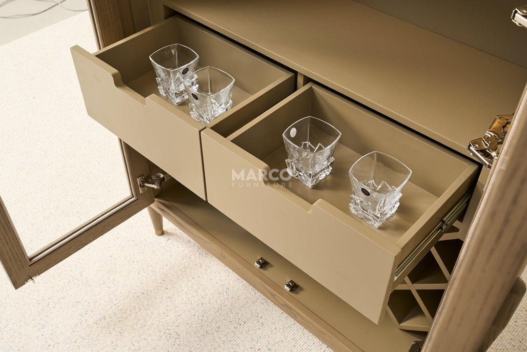 Beige bar cabinet with open drawers containing clear glassware on a light wooden floor.