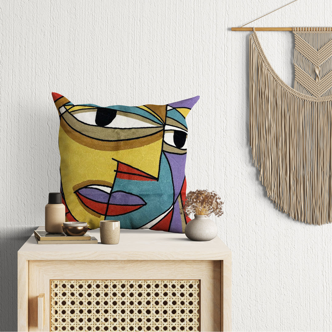 A cushion cover with an abstract multicolored face design, displayed on a wooden side table in a room with a woven wall hanging.