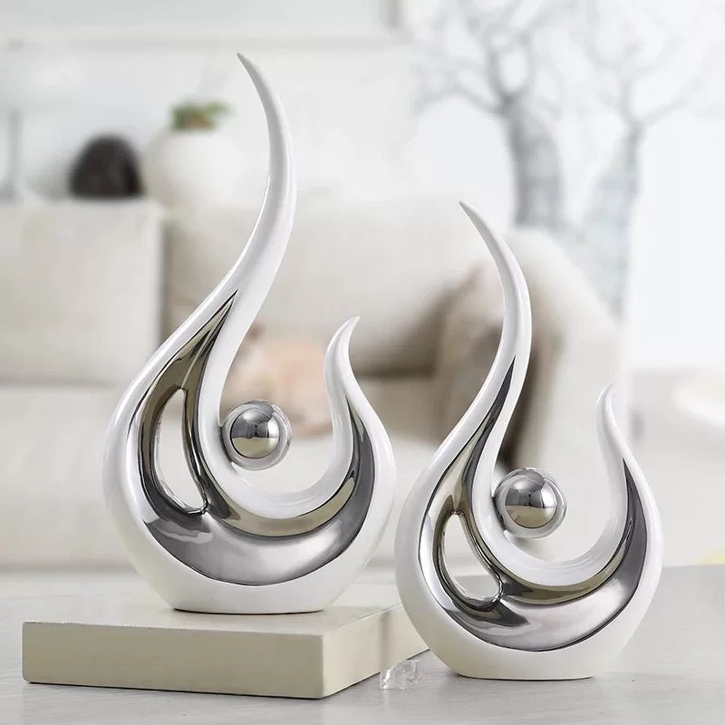 Two modern abstract ceramic phoenix figurines in white and silver colors, displayed on a shelf in a living room setting.